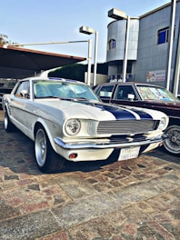 A vintage white car with black racing stripes is parked on a cobblestone surface. The car features classic design elements such as round headlights and chrome accents. It is positioned beside another vehicle and set against a backdrop of a modern building with Arabic signage.