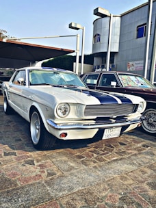 A vintage white car with black racing stripes is parked on a cobblestone surface. The car features classic design elements such as round headlights and chrome accents. It is positioned beside another vehicle and set against a backdrop of a modern building with Arabic signage.