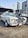 A vintage white car with black racing stripes is parked on a cobblestone surface. The car features classic design elements such as round headlights and chrome accents. It is positioned beside another vehicle and set against a backdrop of a modern building with Arabic signage.