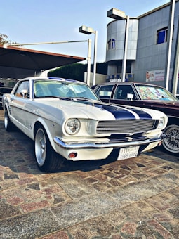 A vintage white car with black racing stripes is parked on a cobblestone surface. The car features classic design elements such as round headlights and chrome accents. It is positioned beside another vehicle and set against a backdrop of a modern building with Arabic signage.