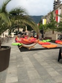 An outdoor seating area is set up with colorful bean bags and a hammock under palm trees on a paved square. The space is surrounded by buildings with balconies draped in banners. A bookshelf is visible in the background, next to dining tables with sun umbrellas.