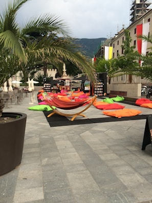 An outdoor seating area is set up with colorful bean bags and a hammock under palm trees on a paved square. The space is surrounded by buildings with balconies draped in banners. A bookshelf is visible in the background, next to dining tables with sun umbrellas.