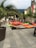 An outdoor seating area is set up with colorful bean bags and a hammock under palm trees on a paved square. The space is surrounded by buildings with balconies draped in banners. A bookshelf is visible in the background, next to dining tables with sun umbrellas.