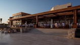 A modern outdoor dining area features wooden architecture with expansive glass windows. Groups of people are seated at tables enjoying an open-air meal as daylight fades. The scene is inviting, with a relaxed atmosphere.