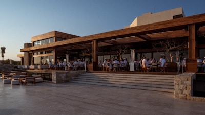 A modern outdoor dining area features wooden architecture with expansive glass windows. Groups of people are seated at tables enjoying an open-air meal as daylight fades. The scene is inviting, with a relaxed atmosphere.