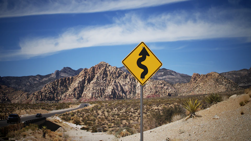 The image depicts a desert landscape with a winding road cutting through it, flanked by red rock formations on both sides. A yellow diamond-shaped sign with black text warning drivers to "curve ahead" is prominently displayed at the center of the scene. In the distance, two cars can be seen driving along the road, and beyond them, majestic mountains rise against a clear blue sky.