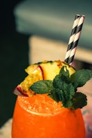 A vibrant orange beverage filled with ice is garnished with fresh mint leaves, a slice of pineapple, and a piece of passion fruit. Two black and white striped straws are inserted into the drink. The background is softly blurred, suggesting a relaxed setting.