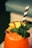 A vibrant orange beverage filled with ice is garnished with fresh mint leaves, a slice of pineapple, and a piece of passion fruit. Two black and white striped straws are inserted into the drink. The background is softly blurred, suggesting a relaxed setting.