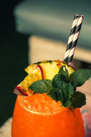 A vibrant orange beverage filled with ice is garnished with fresh mint leaves, a slice of pineapple, and a piece of passion fruit. Two black and white striped straws are inserted into the drink. The background is softly blurred, suggesting a relaxed setting.