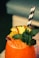 A vibrant orange beverage filled with ice is garnished with fresh mint leaves, a slice of pineapple, and a piece of passion fruit. Two black and white striped straws are inserted into the drink. The background is softly blurred, suggesting a relaxed setting.
