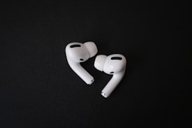 A pair of white, wireless earbuds lie on a dark surface. The earbuds have a sleek and modern design, with visible black accents and silicone ear tips. Their curves and glossy finish stand out against the matte background.
