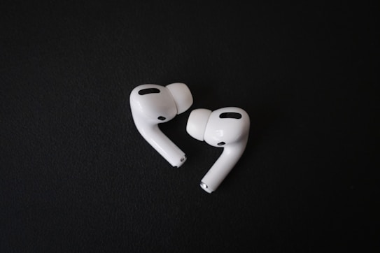 A pair of white, wireless earbuds lie on a dark surface. The earbuds have a sleek and modern design, with visible black accents and silicone ear tips. Their curves and glossy finish stand out against the matte background.