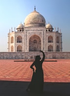 A silhouette of a person striking a pose in front of the Taj Mahal, a majestic white marble mausoleum with detailed decorative patterns and architectural features. The foreground consists of a large, sunlit courtyard with intricate stone paving, contrasting with the clear blue sky in the background.