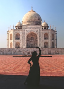 A silhouette of a person striking a pose in front of the Taj Mahal, a majestic white marble mausoleum with detailed decorative patterns and architectural features. The foreground consists of a large, sunlit courtyard with intricate stone paving, contrasting with the clear blue sky in the background.