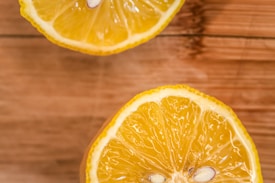 Two lemon halves laid out on a wooden surface. The lemons are cut to reveal the bright yellow, juicy interior with visible seeds. The wooden background adds a natural and rustic feel.