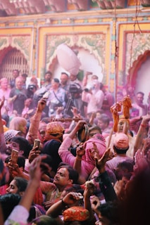 A vibrant crowd celebrates with colorful powders, amidst an ornate backdrop with intricate designs and arches. People are joyfully throwing colored powders in the air, indicating a festive atmosphere.