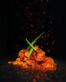 A mound of marinated food, possibly kimchi or a spicy dish, is topped with green garnish and sprinkled with red spices. The dark background contrasts with the vibrant colors of the dish, emphasizing the textures and freshness of the ingredients.