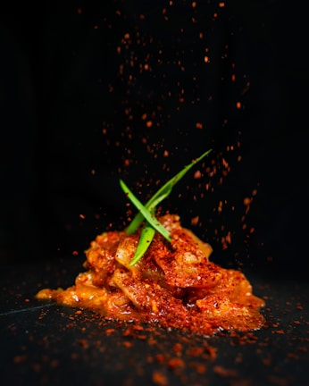 A mound of marinated food, possibly kimchi or a spicy dish, is topped with green garnish and sprinkled with red spices. The dark background contrasts with the vibrant colors of the dish, emphasizing the textures and freshness of the ingredients.