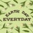 The image features the words 'EARTH DAY EVERYDAY' made from soil or mulch, placed centrally against a pastel green background. Surrounding the text are white tulips with long green leaves, radiating outwards in a pattern.