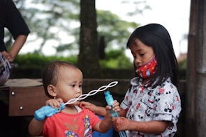 Two children are engaged in play, with a toddler holding a bubble wand and a slightly older child assisting with the bubble mixture. The older child is wearing a colorful mask and has intricate patterns drawn on their hands, suggesting cultural or ceremonial significance. They are outdoors with trees and a wooden structure in the background.