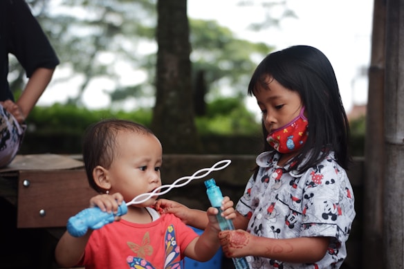 Two children are engaged in play, with a toddler holding a bubble wand and a slightly older child assisting with the bubble mixture. The older child is wearing a colorful mask and has intricate patterns drawn on their hands, suggesting cultural or ceremonial significance. They are outdoors with trees and a wooden structure in the background.