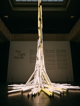 A tall and intricate installation made of numerous fluorescent light tubes that are arranged to create a tower-like structure in the center of a gallery space. The tubes are illuminated, casting a warm glow around the room. The backdrop includes text that reads 'The Light House' on a plain wall, suggesting a theme or title for the exhibit. The ceiling has a grid-like pattern, and the overall atmosphere is modern and artistic.