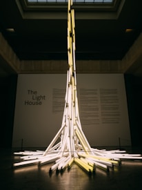 A tall and intricate installation made of numerous fluorescent light tubes that are arranged to create a tower-like structure in the center of a gallery space. The tubes are illuminated, casting a warm glow around the room. The backdrop includes text that reads 'The Light House' on a plain wall, suggesting a theme or title for the exhibit. The ceiling has a grid-like pattern, and the overall atmosphere is modern and artistic.