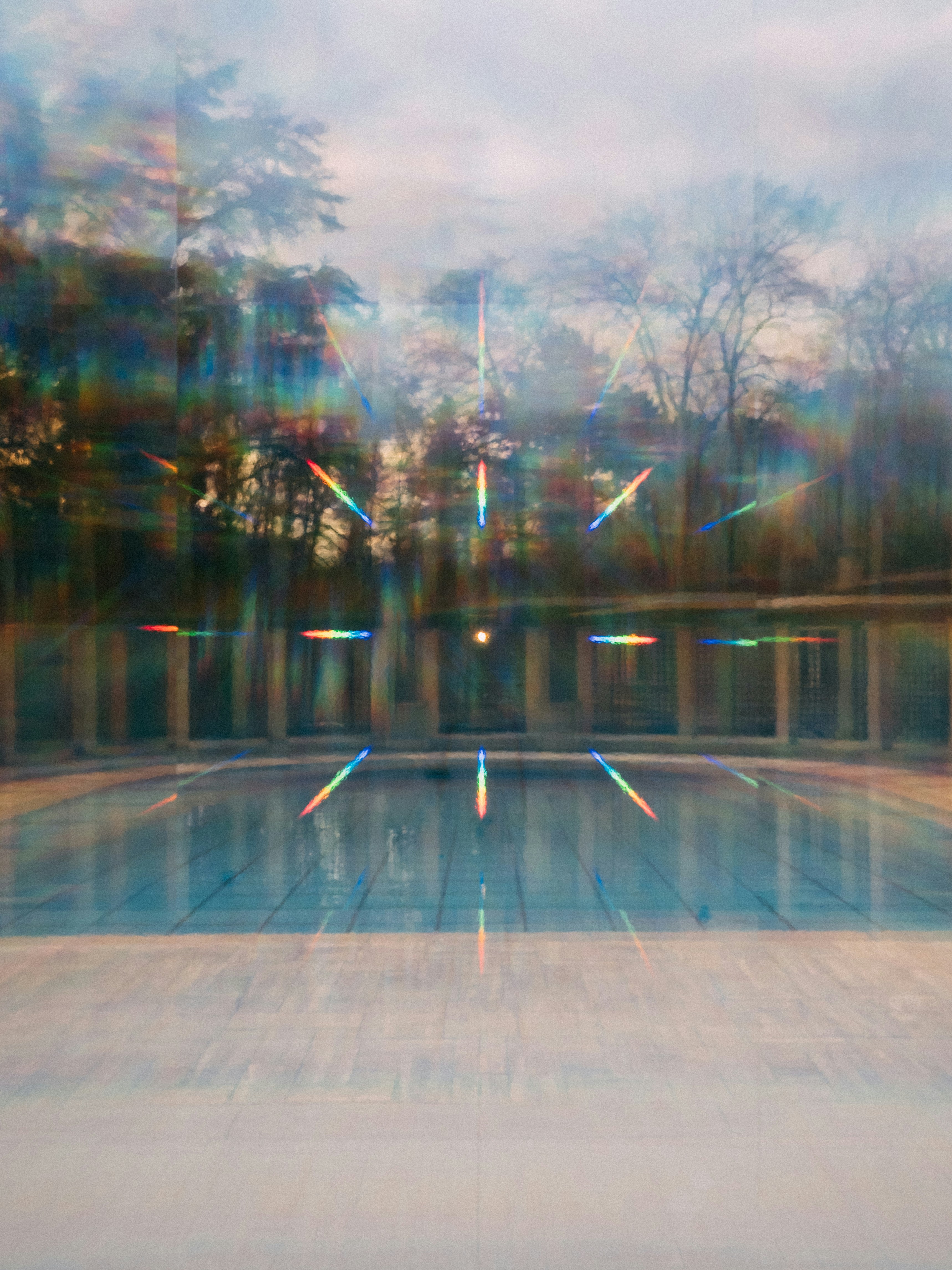 Blurry photo of a blue swimming pool with prismed light and the reflections of trees on a window.
