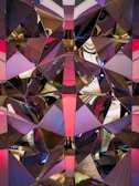 A complex geometric pattern composed of reflective surfaces, creating a kaleidoscopic effect. The surfaces are divided into triangular and polygonal shapes with a mirrored finish, reflecting light and colors in various directions. The reflections include shades of pink, purple, and gold, creating an abstract and dynamic visual experience.