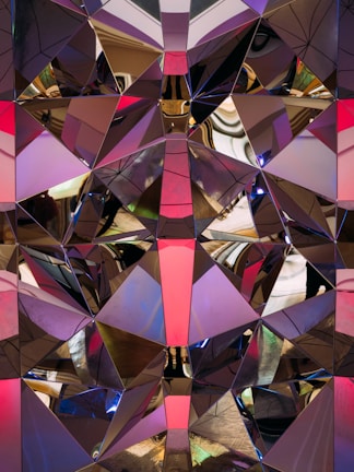 A complex geometric pattern composed of reflective surfaces, creating a kaleidoscopic effect. The surfaces are divided into triangular and polygonal shapes with a mirrored finish, reflecting light and colors in various directions. The reflections include shades of pink, purple, and gold, creating an abstract and dynamic visual experience.