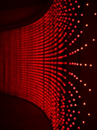 A wall adorned with rows of small, evenly spaced red lights creating a curved, wave-like pattern. The lights appear to be glowing against a dark background, giving a sense of depth and movement.