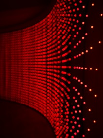 A wall adorned with rows of small, evenly spaced red lights creating a curved, wave-like pattern. The lights appear to be glowing against a dark background, giving a sense of depth and movement.