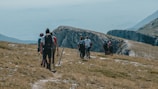 A group of hikers is trekking along a grassy mountain trail. The landscape features rocky outcrops and a distant view of the valley below. The hikers are equipped with backpacks and walking poles, wearing colorful outdoor clothing. The scene is set in a natural, remote, mountainous environment.