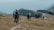 A group of hikers is trekking along a grassy mountain trail. The landscape features rocky outcrops and a distant view of the valley below. The hikers are equipped with backpacks and walking poles, wearing colorful outdoor clothing. The scene is set in a natural, remote, mountainous environment.