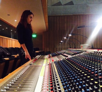 A person is standing in a control room, focused on a large audio mixing console with numerous colorful knobs and sliders. The space features wooden paneling and is illuminated by overhead lights. Seating is visible in the background.