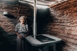 An elderly woman stands inside a rustic, earthen-walled room. She wears a floral blouse and striped skirt, accompanied by a headscarf. The room contains a simple stove with a metal pipe leading upwards, suggesting a traditional or rural setting.