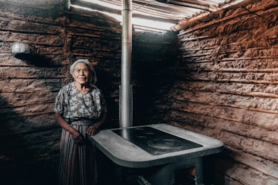 An elderly woman stands inside a rustic, earthen-walled room. She wears a floral blouse and striped skirt, accompanied by a headscarf. The room contains a simple stove with a metal pipe leading upwards, suggesting a traditional or rural setting.