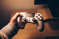 A person is holding a white gaming controller with both analog sticks and various buttons visible. The setting appears cozy, with warm lighting creating a relaxed atmosphere. The background includes a wooden surface, possibly a desk or table.