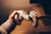 A person is holding a white gaming controller with both analog sticks and various buttons visible. The setting appears cozy, with warm lighting creating a relaxed atmosphere. The background includes a wooden surface, possibly a desk or table.