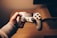 A person is holding a white gaming controller with both analog sticks and various buttons visible. The setting appears cozy, with warm lighting creating a relaxed atmosphere. The background includes a wooden surface, possibly a desk or table.