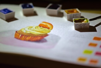 A detailed watercolor painting of a dessert, possibly a cupcake with fruit and cream topping, is on a sheet of paper. Several small watercolor paint pans are scattered around the artwork with a brush dipping into a green hue. Bright color swatches are visible in one corner, suggesting a focus on color mixing and artistic process.