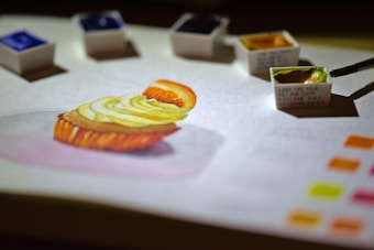 A detailed watercolor painting of a dessert, possibly a cupcake with fruit and cream topping, is on a sheet of paper. Several small watercolor paint pans are scattered around the artwork with a brush dipping into a green hue. Bright color swatches are visible in one corner, suggesting a focus on color mixing and artistic process.