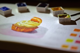 A detailed watercolor painting of a dessert, possibly a cupcake with fruit and cream topping, is on a sheet of paper. Several small watercolor paint pans are scattered around the artwork with a brush dipping into a green hue. Bright color swatches are visible in one corner, suggesting a focus on color mixing and artistic process.