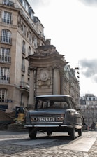 A vintage car with a license plate '643 CI 78' is parked on a cobblestone street in an urban setting. The background features classic European architecture, with ornate buildings and a historical monument or structure. The sky appears overcast, adding a muted tone to the scene.