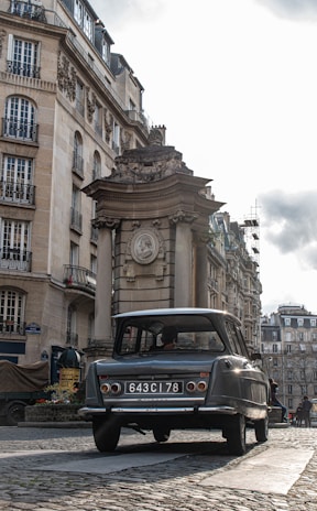 A vintage car with a license plate '643 CI 78' is parked on a cobblestone street in an urban setting. The background features classic European architecture, with ornate buildings and a historical monument or structure. The sky appears overcast, adding a muted tone to the scene.