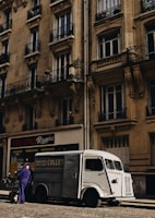 A vintage white delivery van is parked on a cobblestone street in front of a tall, ornate building with multiple windows and balconies. A man wearing a purple uniform is standing next to the van. The building has a sign for 'Studio Reverie' and offers activities like dance, theatre, and music. The overall lighting suggests a late afternoon or early evening ambiance with the street and building slightly shaded.