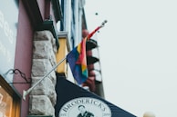 A flag with rainbow colors is attached to the side of a building. Below the flag, there is a sign reading 'Broderick's' with a logo and the establishment year 2019. The building features a mix of stone and brick textures and neighboring structures are visible in the background.