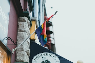 A flag with rainbow colors is attached to the side of a building. Below the flag, there is a sign reading 'Broderick's' with a logo and the establishment year 2019. The building features a mix of stone and brick textures and neighboring structures are visible in the background.
