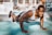 A man wearing a gray tank top is performing push-ups on a turquoise outdoor gym floor. The scene is set in a sunlit outdoor gym with various fitness equipment visible in the background. His expression is focused and determined, and he appears to be in good physical shape.