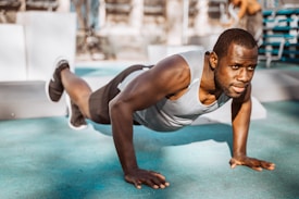 A man wearing a gray tank top is performing push-ups on a turquoise outdoor gym floor. The scene is set in a sunlit outdoor gym with various fitness equipment visible in the background. His expression is focused and determined, and he appears to be in good physical shape.